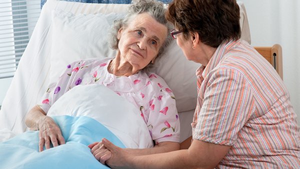 Frau hält die Hand einer älteren Dame, die im Bett liegt