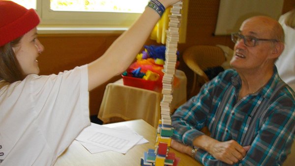 Jugendliche baut mit einem alten Mann einen Turm aus kleinen Holzbausteinen