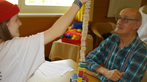 Jugendliche baut mit einem alten Mann einen Turm aus kleinen Holzbausteinen